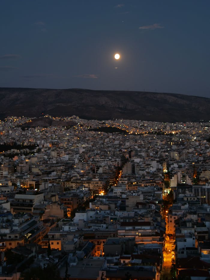 Aerial nighttime view of Athens under a full moon, showcasing city lights and distant mountains.
